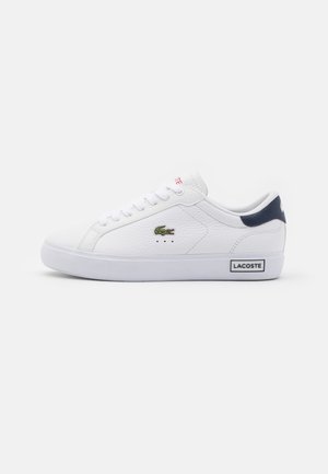 Valkoinen Lacosten matala sneaker, jossa vihreä krokotiililogo sivulla, laivastonsininen kantataki, valkoiset nauhat ja Lacoste-label pohjassa.