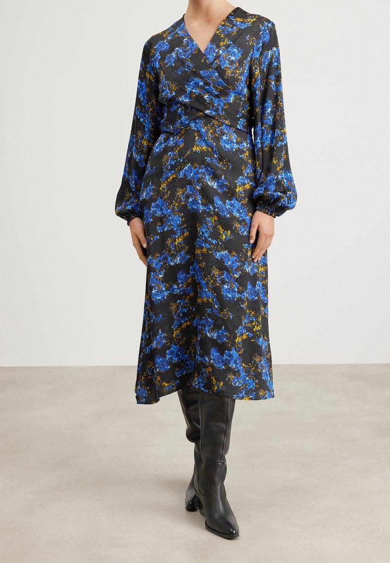 Femme portant une robe midi portefeuille noire à imprimé floral bleu et jaune, associée à des bottes noires montant jusqu'aux genoux, debout sur un sol clair.