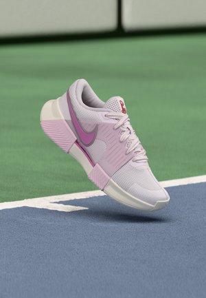 Világoslila és rózsaszín Nike teniszcipő, amely egy kék és zöld teniszpálya felett lóg, fehér határvonalakkal.