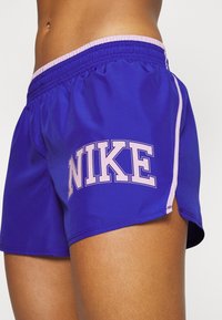 Fialové sportovní šortky vyrobené z hladkého materiálu, s růžovým vnitřním pasem a výrazným bílým logem "NIKE" na boku.