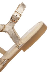 Zlatá kovová plochá sandál s nastavitelným páskem na přezku a polstrovanou béžovou stélkou na světle hnědé gumové podrážce.