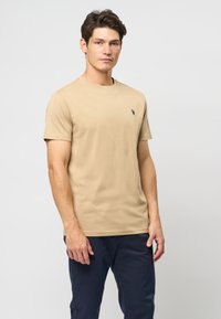 Lysebrun kortærmet t-shirt lavet af bomuld, med et lille sort logo på venstre brystområde. Blød tekstur og klassisk pasform.