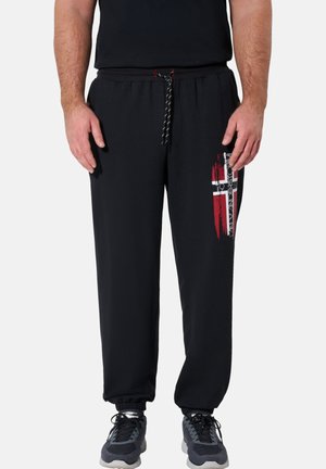 Mann trägt schwarze Jogginghosen mit rotem und weißem Grafikdruck am rechten Bein, schwarzem Kordelzug in der Taille und schwarzen sowie grauen Sneakers.