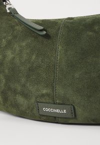 Grønn semsket veske med en glatt overflate, med skinnrem og en merkelapp med "COCCINELLE" i hvit skrift.