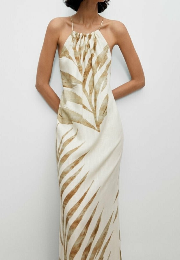 Femme portant une robe longue crème sans manches avec de fines bretelles et de grandes feuilles marron imprimées, debout les bras derrière le dos.