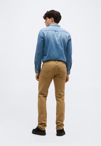 Άνδρας με την πλάτη γυρισμένη, που φοράει μπλε πουκάμισο από τζιν, καφέ τζιν παντελόνι και μαύρα αθλητικά παπούτσια, σε φόντο με απαλό ανοιχτό γκρι χρώμα.