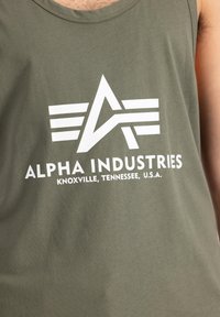 Tricou fără mâneci, de culoare verde măslin, fabricat dintr-un material moale, având un logo mare alb cu accente geometrice și textul "ALPHA INDUSTRIES, KNOXVILLE, TENNESSEE, U.S.A."