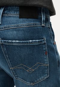 Πίσω τσέπη από μπλε τζιν με χαρακτηριστική μπεζ ραφή ζιγκ-ζαγκ, κόκκινη λεπτομέρεια λογότυπου και μαύρη ετικέτα της μάρκας πάνω από το τελείωμα σκούρου γκρι πουκαμίσου.