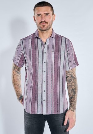 Man with short hair and tattoos wearing a short-sleeve, button-up shirt with vertical purple, gray, and white stripes.