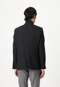 Black tailored blazer with a high collar and single vent at the back, paired with patterned gray trousers featuring a geometric design.