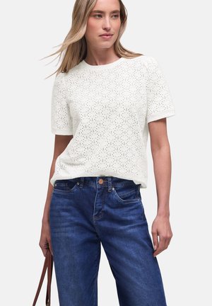 Kvinde iført en hvid kortærmet perforeret top og blå jeans, der holder en brun håndtaskestropp i venstre hånd.