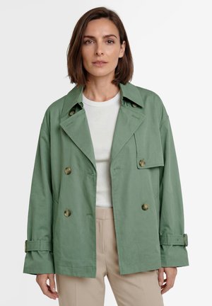 Mujer con cabello castaño hasta los hombros que lleva una chaqueta verde claro de doble botonadura, camiseta blanca y pantalones beige sobre un fondo liso.
