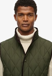 Gilet matelassé vert olive avec des détails en brun, doté d'un col haut et d'une fermeture par pression, porté par-dessus un pull en laine crème.