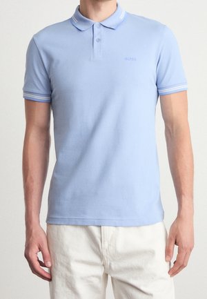 Homme portant un polo bleu clair à manches courtes avec des rayures blanches sur le col et les manches, associé à un pantalon écru.