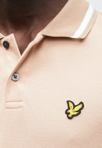 Világos barack színű pólóing texturált anyagból, fekete gombokkal és sárga hímzett madár logóval a bal mellrészen.