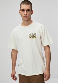 Młody mężczyzna z krótkimi włosami, ubrany w biały t-shirt z czarnym i żółtym napisem graficznym "Coalition" oraz brązowe spodnie, na jednolitym tle.