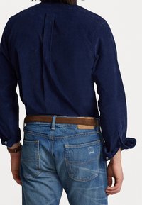 Mörkblå kord skjorta med vikta ärmar, kombinerad med ljusblå denimjeans med distinkta bakfickor och ett brunt läderbälte.