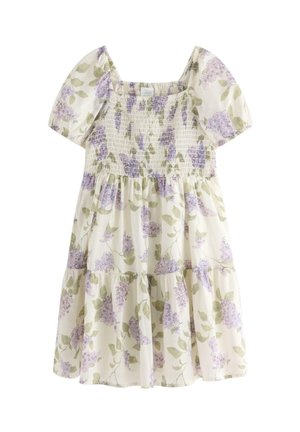 Smilende ung pige i en blomsterkjole, hvid strikket cardigan, lilla hårbånd, lavendel sokker og beige sko står på ét ben.