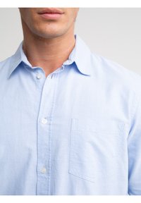 Mann trägt ein hellblaues Hemd mit Knöpfen, Kragen und Brusttasche, zeigt unteren Teil des Gesichts und den Hals vor einem einfarbigen Hintergrund.