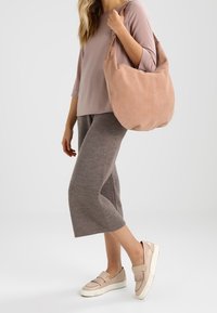 Světle růžová semišová taška přes rameno s jemným, zakřiveným tvarem. Nosit s volným růžovým topem, texturovanými béžovými culotami a neutrálními plochými botami.