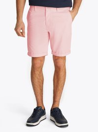 Homem vestido com calções rosa arregaçados e ténis pretos, de pé com uma mão no bolso contra um fundo branco.