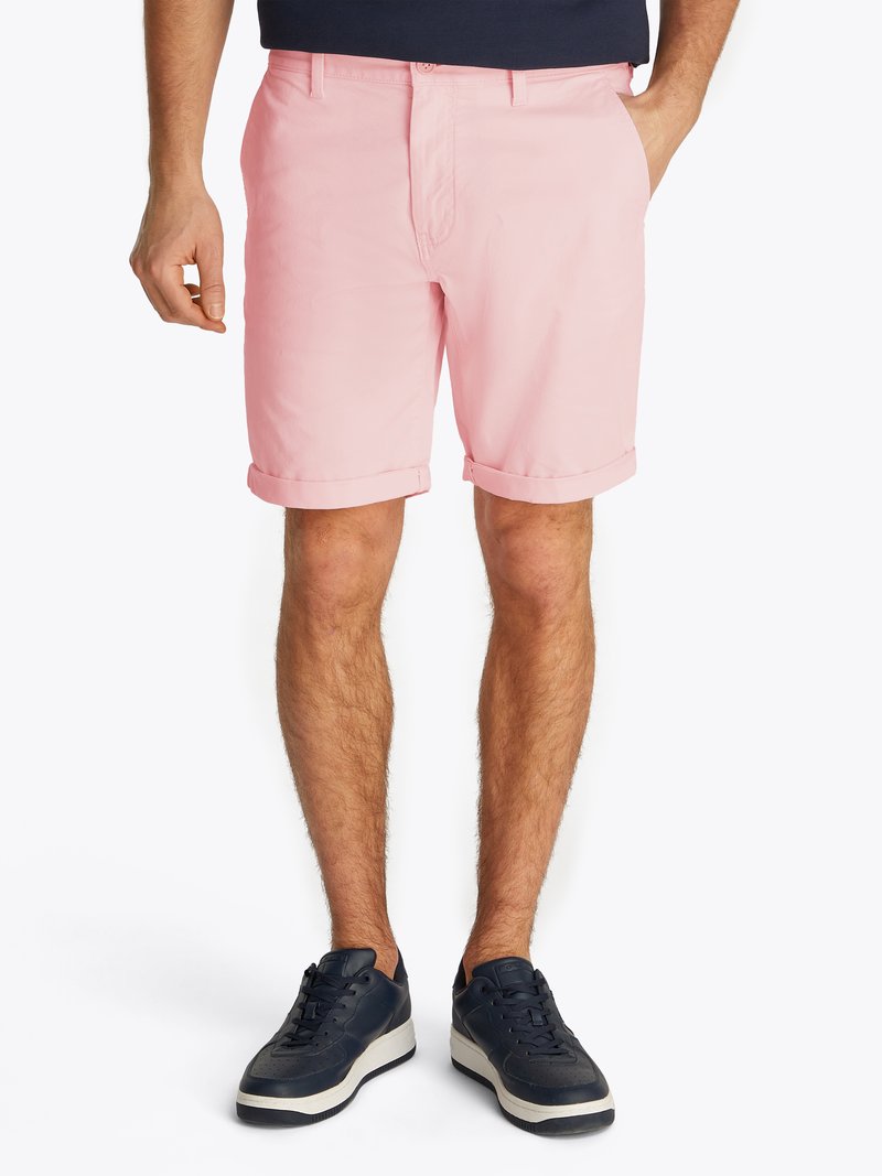 Homem vestido com calções rosa arregaçados e ténis pretos, de pé com uma mão no bolso contra um fundo branco.