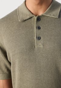 Gros plan d'une personne portant un polo en maille vert olive à manches courtes avec un col et trois boutons foncés.