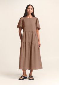 Vestito marrone di lunghezza midi con maniche a sbuffo corte, scollatura rotonda e una gonna a strati. Indossato con sandali neri, su uno sfondo neutro.