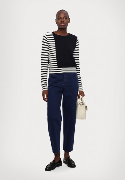 Strukturert genser med svart og hvitt stripemønster, designet som en cropped. Kombinert med mørkeblå bukser. Modellen holder en liten kremfarget håndveske. Svarte loafers fullfører antrekket.