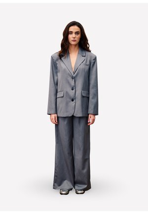 Vrouw met lang, donker haar, gekleed in een oversized grijze suit met wijde broekspijpen, staand en recht vooruit kijkend tegen een plain witte achtergrond.