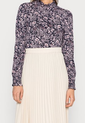 Kobieta ubrana w czarną bluzkę z długim rękawem i różowym kwiatowym wzorem oraz z wysokim kołnierzem, do tego biała plisowana spódnica z wysokim stanem.