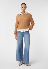 Frau, die einen beigen Strickpullover über einem weißen Kragenhemd, weite blaue Jeans und beige Absätze trägt.