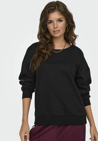 Zwarte sweatshirt met een losse pasvorm, ronde halslijn en verlaagde schouders. Zachte stof, ribbelboorden en onderzoom. Gecombineerd met bordeauxrode broek.