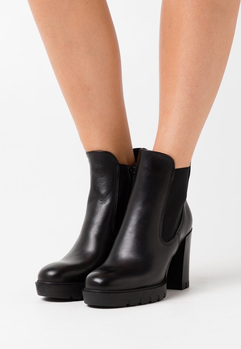 Bottes noires en cuir à talons hauts épais portées sur des jambes nues sur un fond blanc.