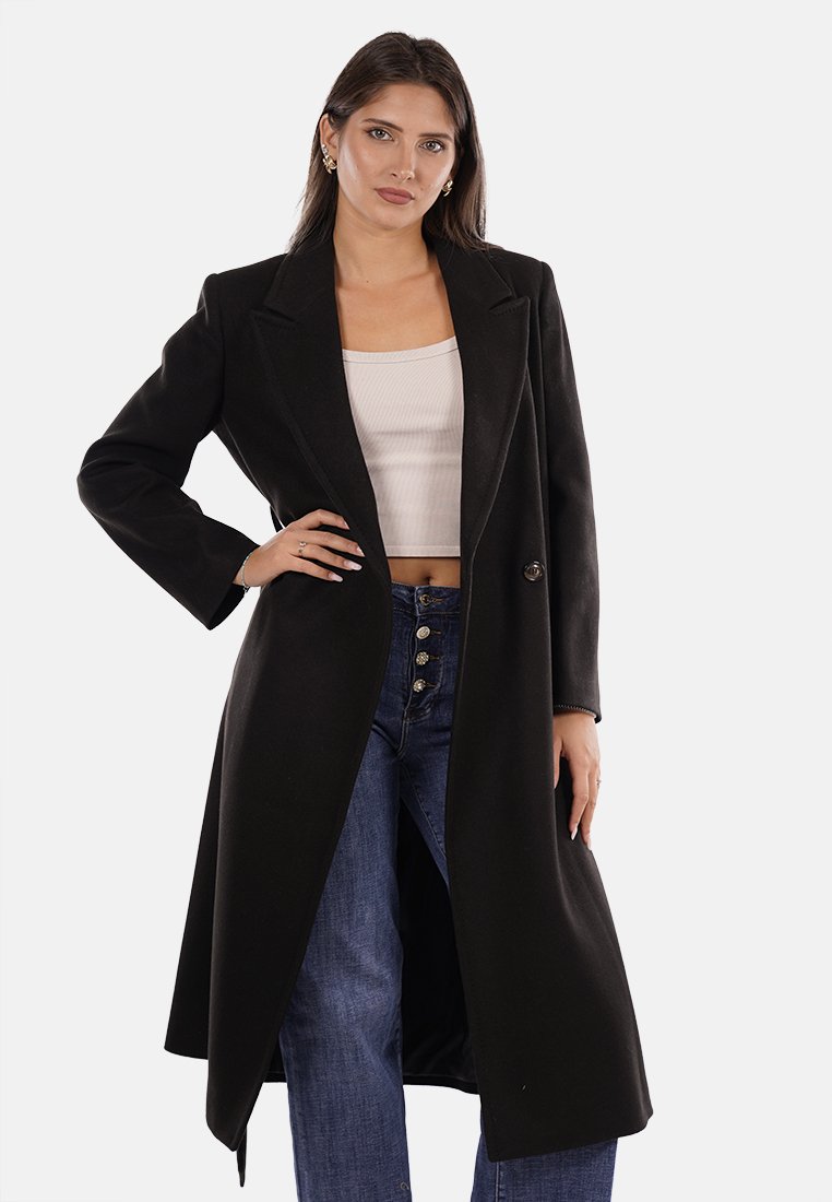 Donna con lungo cappotto nero, top bianco a maniche corte e jeans blu a vita alta, in piedi con una mano sui fianchi su uno sfondo bianco.