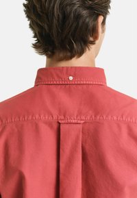 Rotes Baumwollhemd mit Kentkragen, Rückenfalte und einem einzelnen Knopfdetail am Hals. Glatte Textur ohne sichtbare Muster.