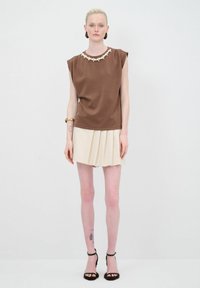 Top marrón de manga corta con un escote decorativo de cuentas blancas, combinado con una mini falda plisada color crema y sandalias de tacón negro.
