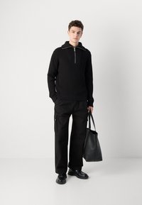 Suéter negro de canalé con cuello de media cremallera, combinado con pantalones cargo negros holgados. El modelo sostiene un bolso de mano de cuero negro. Textura suave.