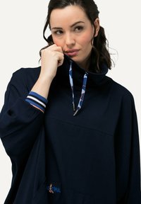 Merinovillane pullovere kõrge kraega, kuldsete detailide, sinise ja oranži mustriga nööriga ning triibuliste varrukatega. Ees on tasku, millel on tekst.