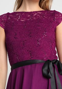 Robe en dentelle violette à manches courtes, décorée de petits sequins, avec un ruban en satin noir noué en un nœud à la taille.