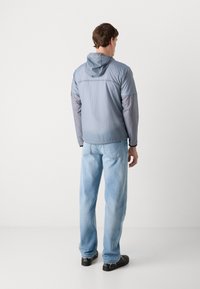 Lightweight, hooded jacket in a muted blue tone, featuring a smooth texture and contrasting gray sleeves. Paired with light blue jeans and black shoes.