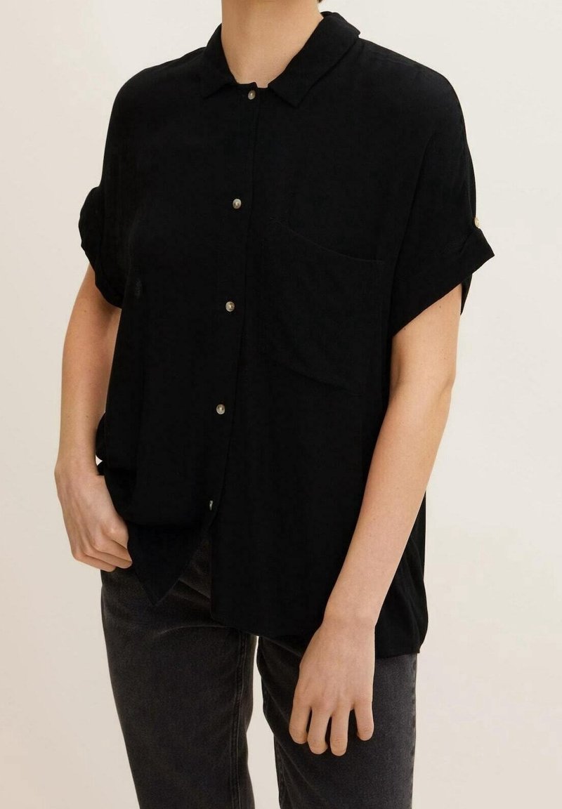 Personne portant une chemise noire à manches courtes avec boutons, col et poche avant, associée à un pantalon gris foncé.