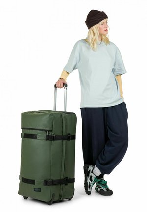 Person trägt eine schwarze Mütze, hellblaues Hemd und marineblaue Hose und lehnt sich an einen großen grünen Rollkoffer mit ausgezogenem Griff.