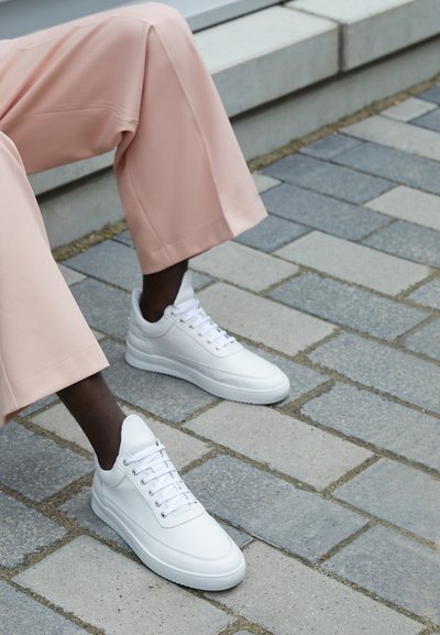 White leather sneakers with a smooth texture, round toe, and lace closure, paired with light pink, wide-legged trousers on a stone pavement.