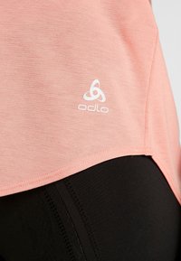 Růžové sportovní tričko Odlo s hladkou texturou, kulatým zakončením a bílým logem. Kombinováno s černými sportovními legínami.