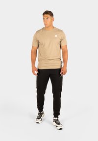 Beige t-shirt met korte mouwen en een subtiele logo op de borst, gecombineerd met zwarte joggerbroeken met zijstrepen en zakken.