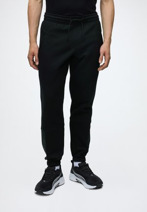 Hombre con pantalones deportivos negros con cintura de cordón, calcetines blancos y zapatos deportivos en blanco y negro, de pie sobre fondo liso.