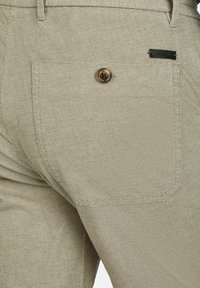 Beige patroonbroek van een gestructureerde stof, met een achterzak met een ronde bruine knop en een klein zwart label eraan bevestigd.