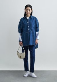 Τουνίκ από τζιν με μπλε και λευκές ριγέ μανίκια, λεπτομέρεια με κουμπιά, συνδυασμένη με μπλε παντελόνι και λευκά αθλητικά παπούτσια, κρατώντας μια κρεμ τσάντα.