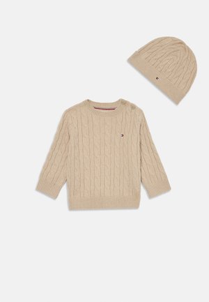 Pulôver bege com ponto de cable, de mangas longas e fecho lateral em botão, combinado com um gorro canelado a condizer, ambos em textura de malha suave.